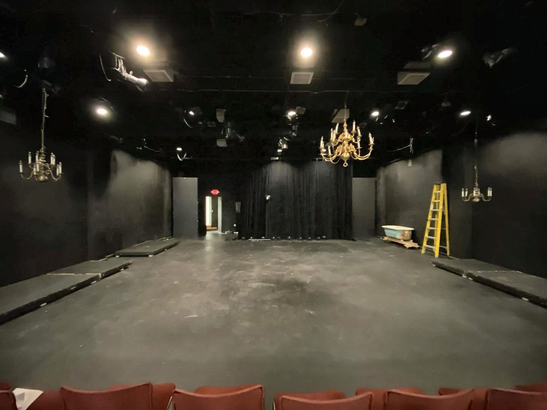 Jones 125 empty: an open black floor with black walls and curtains, stage lights and chandeliers overhead, seen from the front row