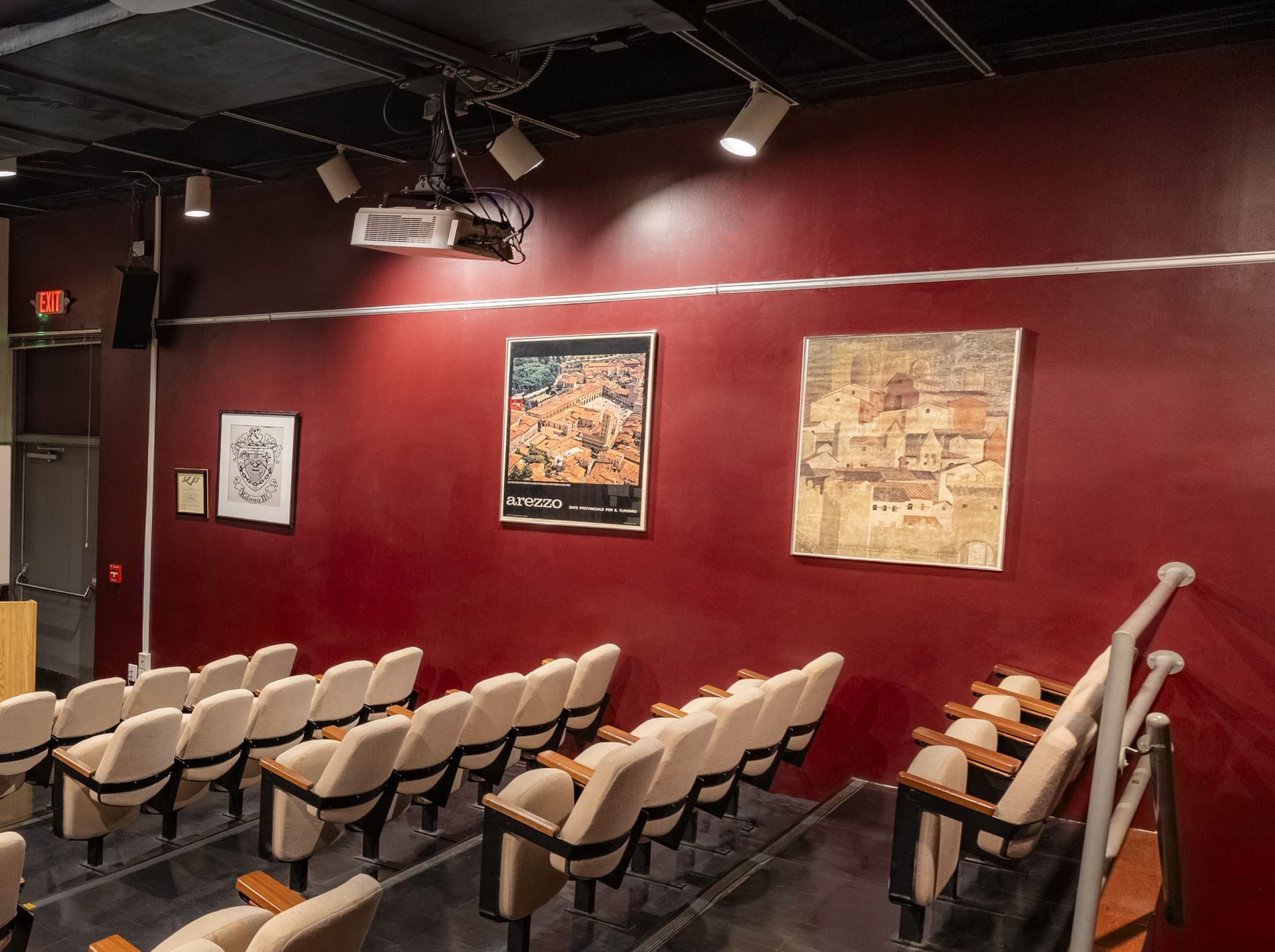 Ceiling-mounted projector and spotlights above tiered seating and framed posters on a red wall in Jones 125